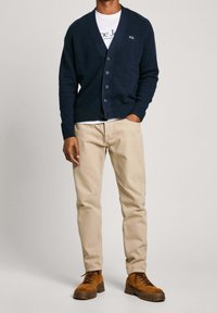 Laivastonsininen neuletakki V-pääntiellä, napillinen edusta, resoriset hihansuut ja kirjailtu logo. Beige suorat housut ja ruskeat saappaat.