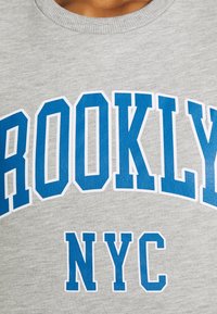 Gros plan d'un sweat-shirt gris avec des lettres bleues en bloc formant "BROOKLYN" au-dessus de "NYC" sur le devant.