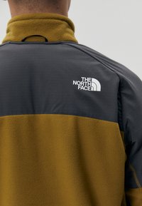 Giacca in fleece con una parte superiore grigia e una parte inferiore giallo senape. Caratterizzata da un colletto alto e un logo bianco sul retro.