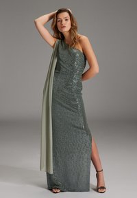 Ein trägerloses Abendkleid aus dunkelgrünen Pailletten mit drapiertem, hellgrünem Stoff; verfügt über einen seitlichen Schlitz und eine figurbetonte Silhouette. Getragen mit schwarzen Sandalen.