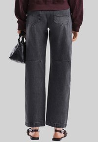 Jeans a gamba larga a vita alta in denim grigio scuro, con due tasche posteriori e dettagli di cucitura discreti, abbinati a una borsa nera e sandali.