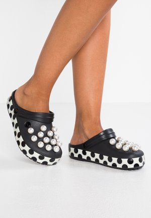 Sabots noirs avec semelles à pois blancs et grandes décorations blanches en forme de perles, portés pieds nus sur fond blanc.