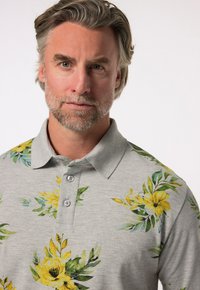 Grå polotröja med ett blommönster som har gula blommor och gröna blad. Den har en klassisk krage och en knäppning med tre knappar.