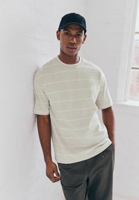 Light green short-sleeve shirt with white stripes, paired with dark pants. The model stands against a white wall. Black cap worn.