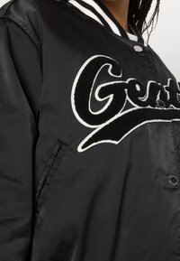 Sort satin varsityjakke med et hvidt broderet logo, stribet krave og trykknaplukning; glat tekstur, sidelommer.