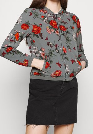 Kvinde iført en grå lynlåstrøje med rødt og pink blomstermønster og sort denimnederdel med frynset kant, hænderne i jakke-lommerne.