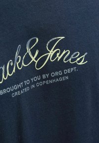 Tricou din bumbac de culoare bleumarin, cu text imprimat alb și verde deschis: "Jack & Jones" și detalii suplimentare de design dedesubt.