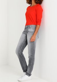 Rood langarm shirt, grijze skinny jeans en witte sneakers. De outfit is eenvoudig met een aansluitend ontwerp en zachte texturen. Geen opmerkelijke patronen.