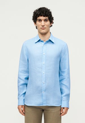 Tânăr cu păr închis la culoare și creț, purtând o cămașă bleu deschis cu nasturi și pantaloni bej, stând în fața unui fundal simplu, deschis la culoare.