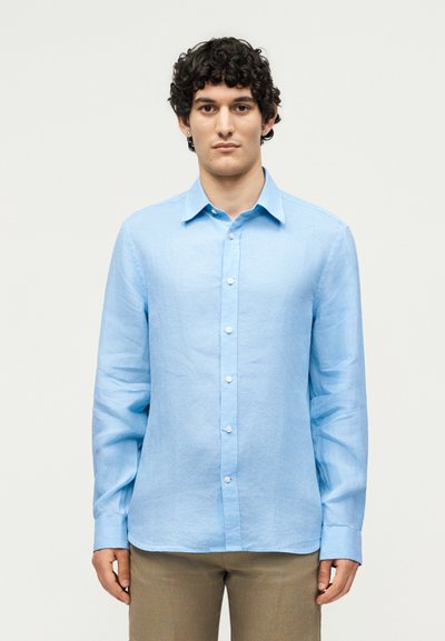 Joven con cabello rizado oscuro, vestido con una camisa azul claro abotonada y pantalones beige, de pie frente a un fondo claro y simple.