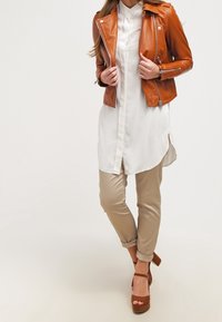 Brązowa skórzana kurtka motocyklowa na białej koszuli zapinanej na guziki z asymetrycznym dołem, w połączeniu z beżowymi spodniami i brązowymi platformami na obcasie.