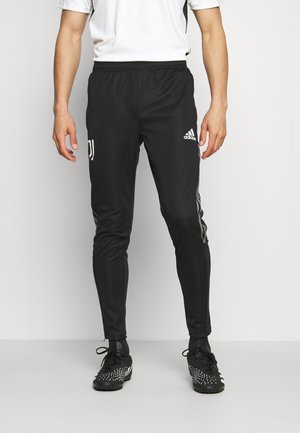 Un homme portant un pantalon de soccer Adidas Juventus noir et des crampons noirs, debout devant un fond clair uni.