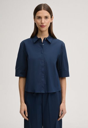 Frau mit glatten braunen Haaren, die ein marineblaues, kurzärmeliges Hemd mit Knopfleiste und passende Hose trägt, stehend vor einem einfarbigen Hintergrund.