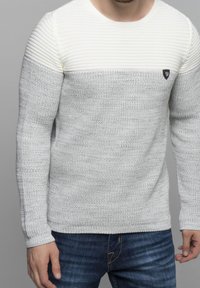 Pull en maille gris clair avec un panneau texturé blanc en haut, comportant un petit logo noir sur la poitrine. Encolure ronde et manches longues.