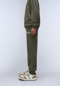 Olivengrüner Sweatshirt und passende Jogginghose in lässiger Passform, mit gerippten Bündchen und einem logo in passendem Farbton. Ein Paar weiße und beige Sneaker.