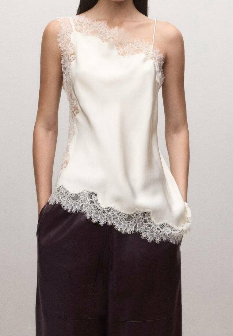 Femme portant un caraco en satin blanc orné de dentelle avec des bretelles fines et un pantalon ample foncé, debout les mains dans les poches.