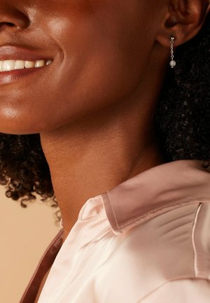 Silberne Ohrhängerteile mit einem weißen Perlenakzent, getragen von einer Person mit lockigem Haar, kombiniert mit einer blassrosa Satinbluse.