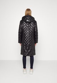 Cappotto lungo nero trapuntato con una texture lucida, cappuccio e motivo a diamante. Indossato con pantaloni scuri e sneaker bianche.