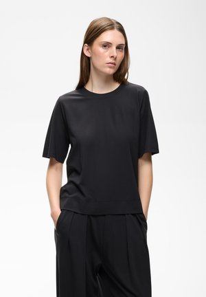 Femme aux cheveux bruns raides portant un t-shirt noir uni à manches courtes et un pantalon noir, les mains dans les poches, debout devant un fond blanc.