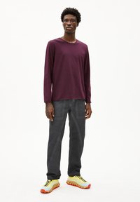 Lange mouwen paarse T-shirt, grijze rechttoe rechtaan broek en gele sneakers met rode zolen. Eenvoudig ontwerp, zacht materiaal en contrasterende kleuren.