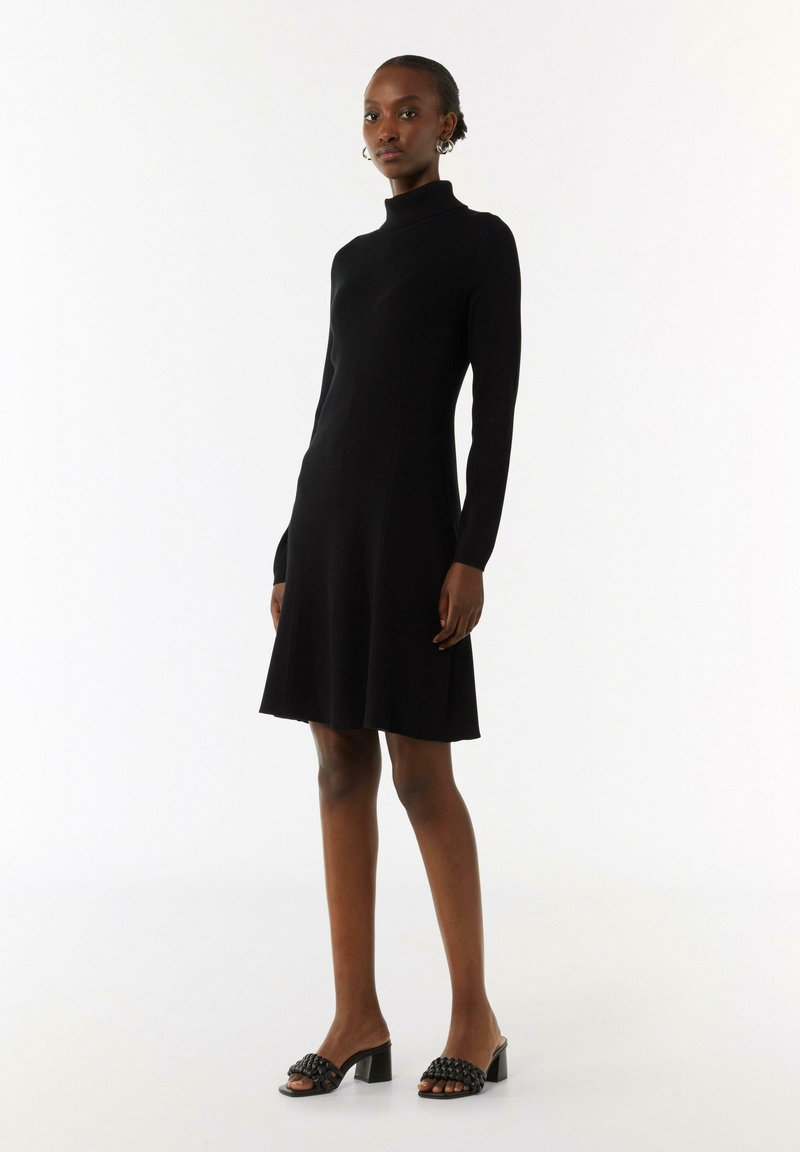 Schwarzes geripptes Strickkleid mit einem figurnahen Rollkragen und langen Ärmeln, das am Saum leicht ausgestellt ist. Kombiniert mit schwarzen Absatzmules.