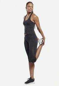 Naine mustas spordivarustuses venitab ühte jalga taha, hoides jalast kinni oma käega, vaatab kõrvale ja naeratab.