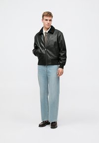Černá kožená bomber bunda s texturovaným kožešinovým límcem, se zipovým zapínáním a ribbed manžetami, v kombinaci s světle modrými džíny rovného střihu a botami.