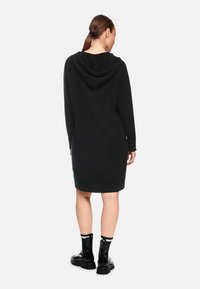 Schwarzes Kapuzenkleid mit geripptem Saum und Bündchen, aus einem weichen Material gefertigt. Getragen mit glänzenden schwarzen Knöchelstiefeln und Socken mit weißen Akzenten.