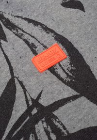 Grijze stof met een zwart bladmotief, met een gestructureerd oranje label waarop "U&JOY" duidelijk in de stof is gestikt.