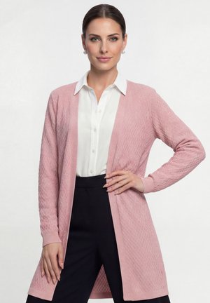 Žena s uhlazenými vlasy staženými dozadu, nosící perlové náušnice, bílou košili s límečkem, růžový texturovaný svetr a černé kalhoty, stojící s jednou rukou na boku.