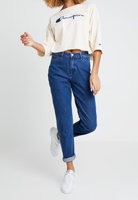 Weißer, kurzer Sweatshirt mit blauer, gestickter "Champion"-Logos, kombiniert mit hoch geschnittenen blauen Denim-Jeans und weißen Sneakers.