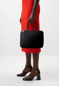 Borsa tote in pelle nera con due manici, chiusura a zip e logo argentato. Indossata con un vestito rosso e tacchi a spillo neri.
