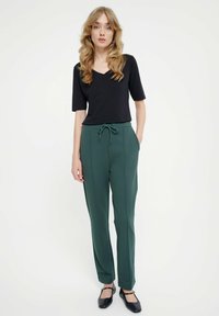 Czarna bluzka z krótkim rękawem i dekoltem w serek, zestawiona z zielonymi, dopasowanymi spodniami z troczkiem w talii, bocznymi kieszeniami i gładką fakturą.