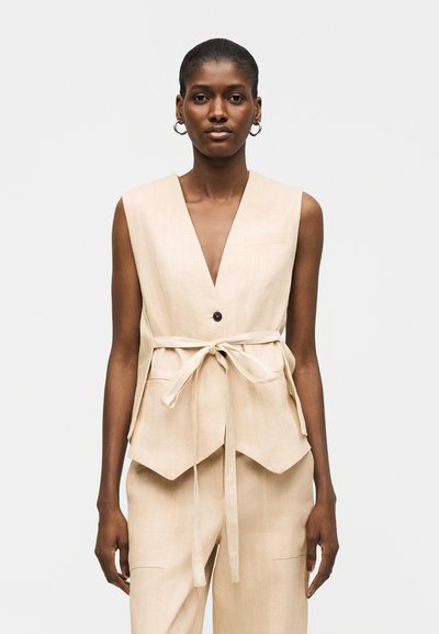 Femme aux cheveux courts portant un gilet sans manches beige ceinturé et un pantalon assorti, debout devant un fond clair uni.