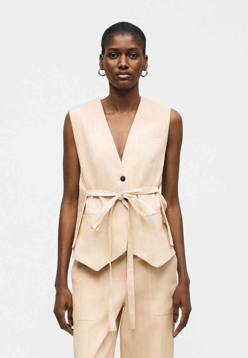 Γυναίκα με κοντά μαλλιά, ντυμένη με μπεζ αμάνικο γιλέκο με ζωνάκι και παντελόνι σετ, στέκεται μπροστά σε ένα απλό φωτεινό φόντο.