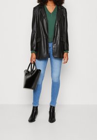 Svart imitationsläder överdimensionerad kavaj över en grön tröja, i kombination med ljusblå tajta jeans och svarta ankelskor. Håller en liten svart handväska.