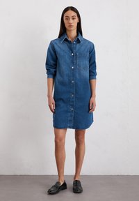 Denim-Hemdkleid in Mittelblau, mit langen Ärmeln, durchgehender Knopfleiste, einer Brusttasche und einem abgerundeten Saum. Getragen mit schwarzen Slippern.