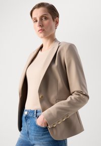 Beige přizpůsobený blazer se zlatými knoflíky a hladkou texturou, nošený přes na ribbovaný světlý top a doplněný modrými džíny.