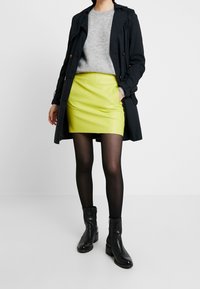 Schwarzer Trenchcoat über einem grauen Pullover, kombiniert mit einem leuchtend gelben Leder-Minirock und schwarzen Stiefeletten; schwarze Strumpfhosen runden den Look ab.