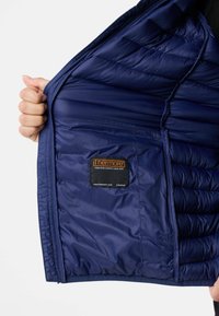 Laivan tummansininen eristetty takki, jossa on tikattu vuori ja Thermaore-merkki. Takki on valmistettu sileästä, kiiltävästä kankaasta ja siinä on vetoketjullinen tasku.