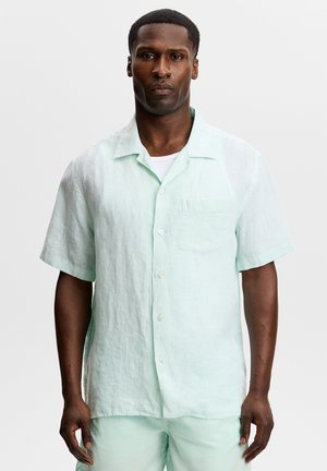 Hombre con expresión seria que lleva una camisa de manga corta verde claro abotonada y pantalones a juego sobre fondo blanco.