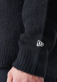 Černý pletený svetr s rukávem s ribbováním. Obsahuje malou bílou nášivku poblíž manžety. Měkký, huňatý materiál.