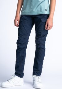 Námornické cargo kalhoty s mnoha bočními kapsami, vyrobené z odolného bavlněného materiálu, s úzkým střihem a jemnou texturou. Na nohou bílé tenisky.