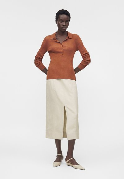 Γυναίκα που φοράει μακρυμάνικη ριπ ρουχιστική πόλο σε σκουριά απόχρωση, ανοιχτόχρωμη μίντι φούστα με σκίσιμο μπροστά και μπεζ μπαλαρίνες με μυτερή μύτη και λουράκια στον αστράγαλο.