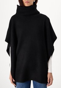 Poncho noir en tricot avec un col roulé haut. Présente des côtes texturées, une coupe ample et des ouvertures latérales. Associé à un jean bleu.