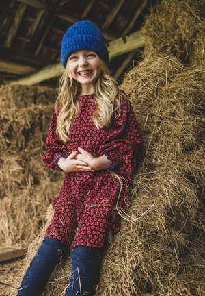 Kind dat een rode bloemenjurk met pofmouwen en blauwe accenten draagt, zittend op hooi balen, glimlachend, met lang golvend blond haar en een blauwe gebreide muts.
