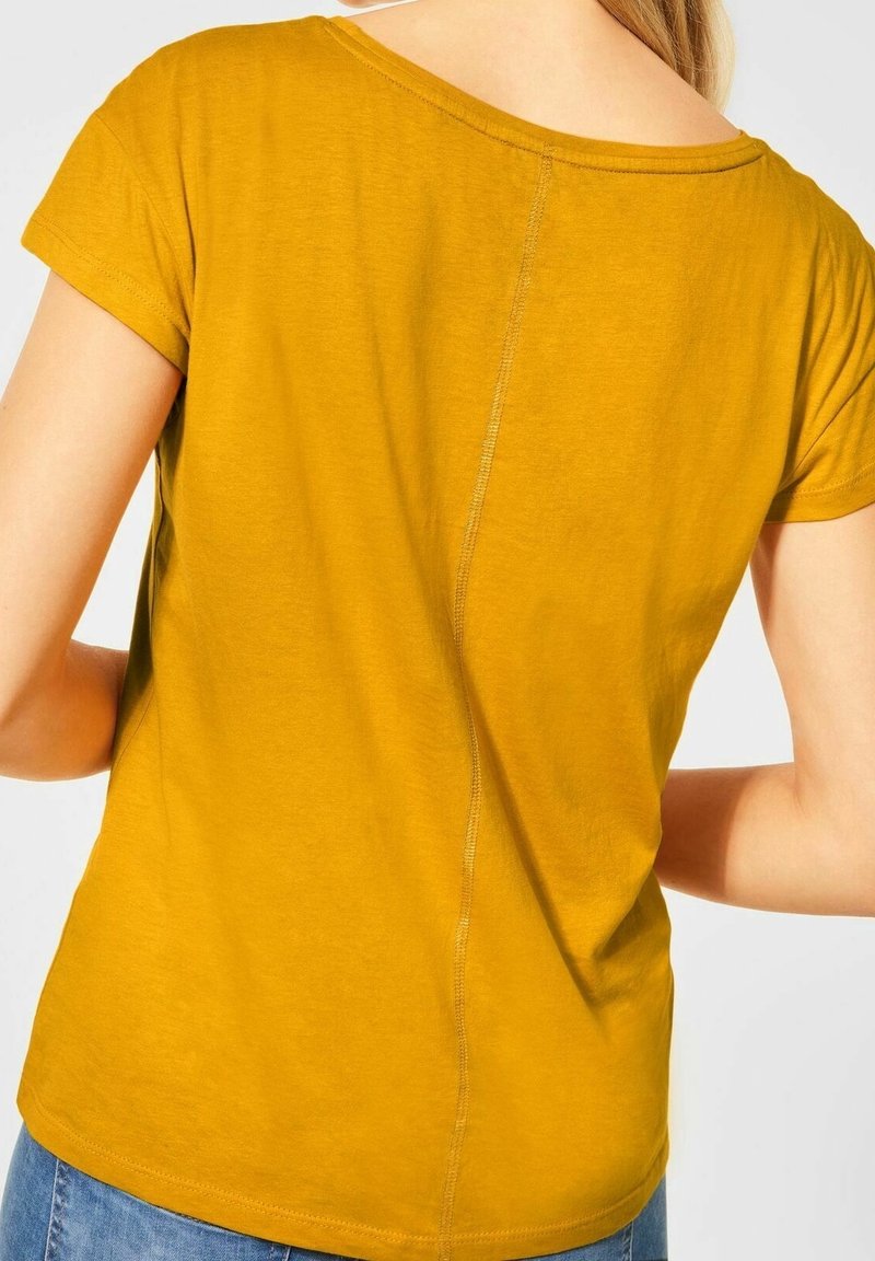 Žluté tričko s krátkým rukávem a kulatým výstřihem, se středovým švem na zadní straně a měkkou texturou. Spodní lem se mírně zakřivuje.