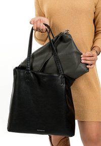 Personne en robe beige tenant un sac fourre-tout en cuir noir avec poignées et une pochette en cuir noir à fermeture éclair sur fond blanc.