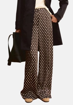 Donna che indossa un cappotto nero, un top bianco, pantaloni marroni a gamba larga con motivo a rombi bianchi, scarpe beige e tiene in mano una borsa nera.