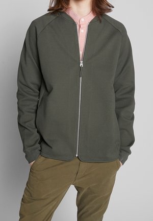 Person trägt eine offene olivgrüne Jacke über einem rosafarbenen Hemd mit Kragen und olivgrüner Hose, die Hände in den Taschen, steht vor einem grauen Hintergrund.